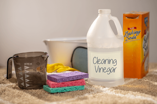 Vinegar Cleaning Bottle of cleaning vinegar, box of baking soda, dish sponges, measuring cup, gloves, and bowl on carpet